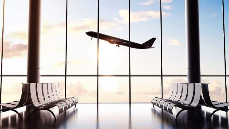 A picture of an airport gate area with empty chairs. Outside of the window, a plane is taking off and backlit by a bright sky.